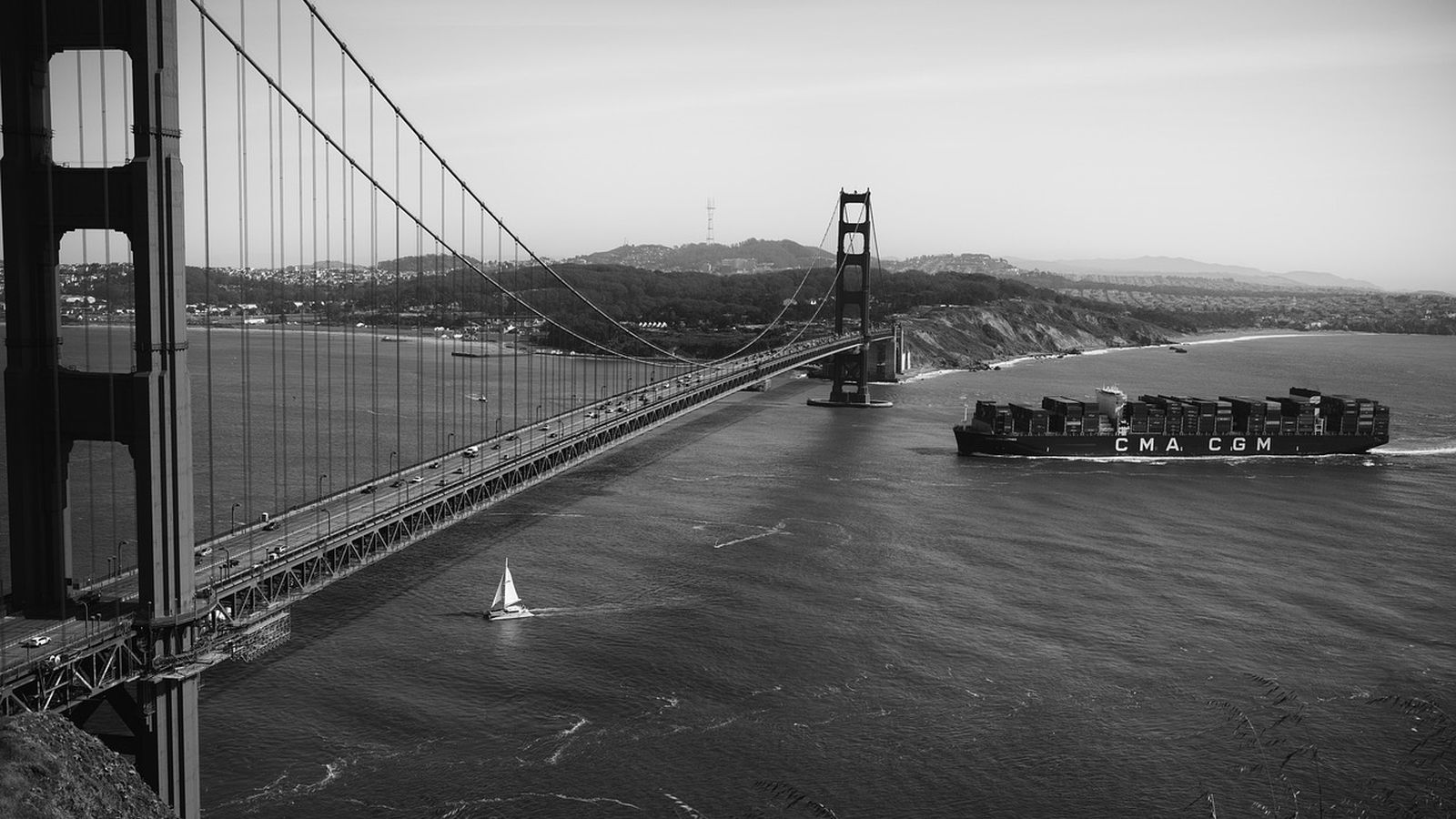 A small sailing boat and a large container ship passing beneath the Golden Gate Bridge